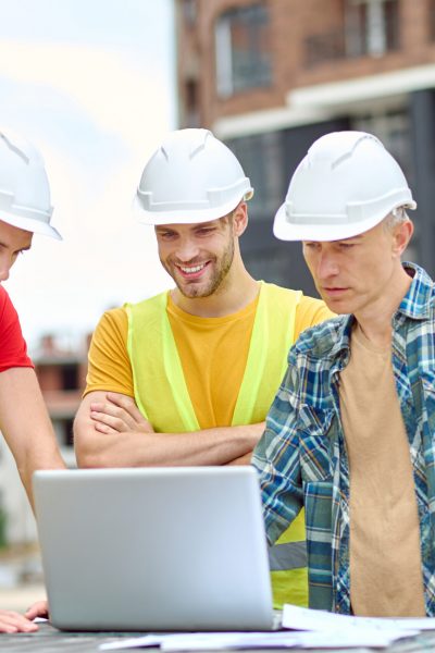 Important information. Three men in protective helmet looking at laptop with interest while standing at construction site during day