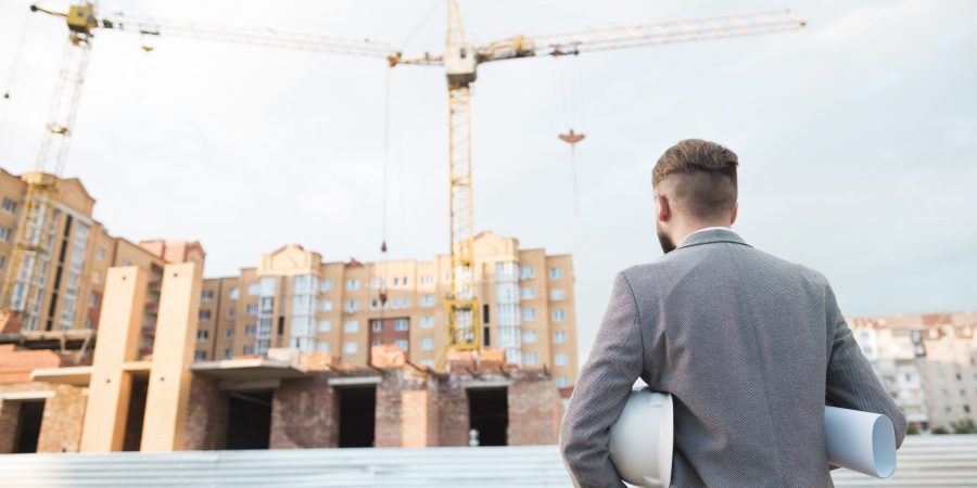 rear-view-male-architect-holding-blueprint-hard-hat-looking-construction-site
