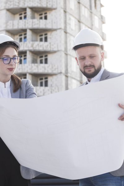 two-professional-architect-holding-blueprint-looking-it-near-construction-site